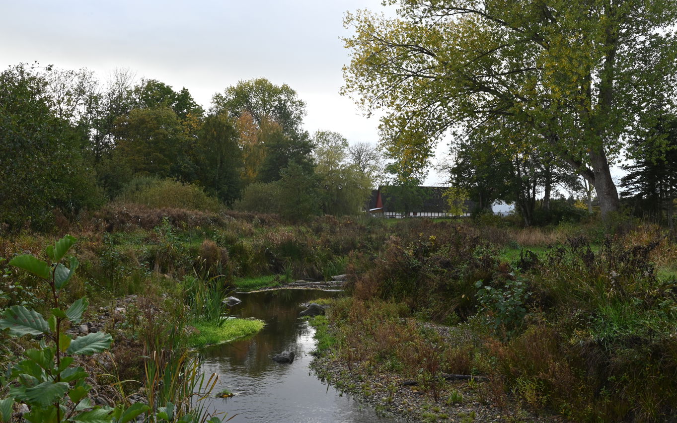I forgrunden et lavvandet vandløb og i baggrunden en rød/hvid bindingsgsværksgård. Mange træer i kanterne