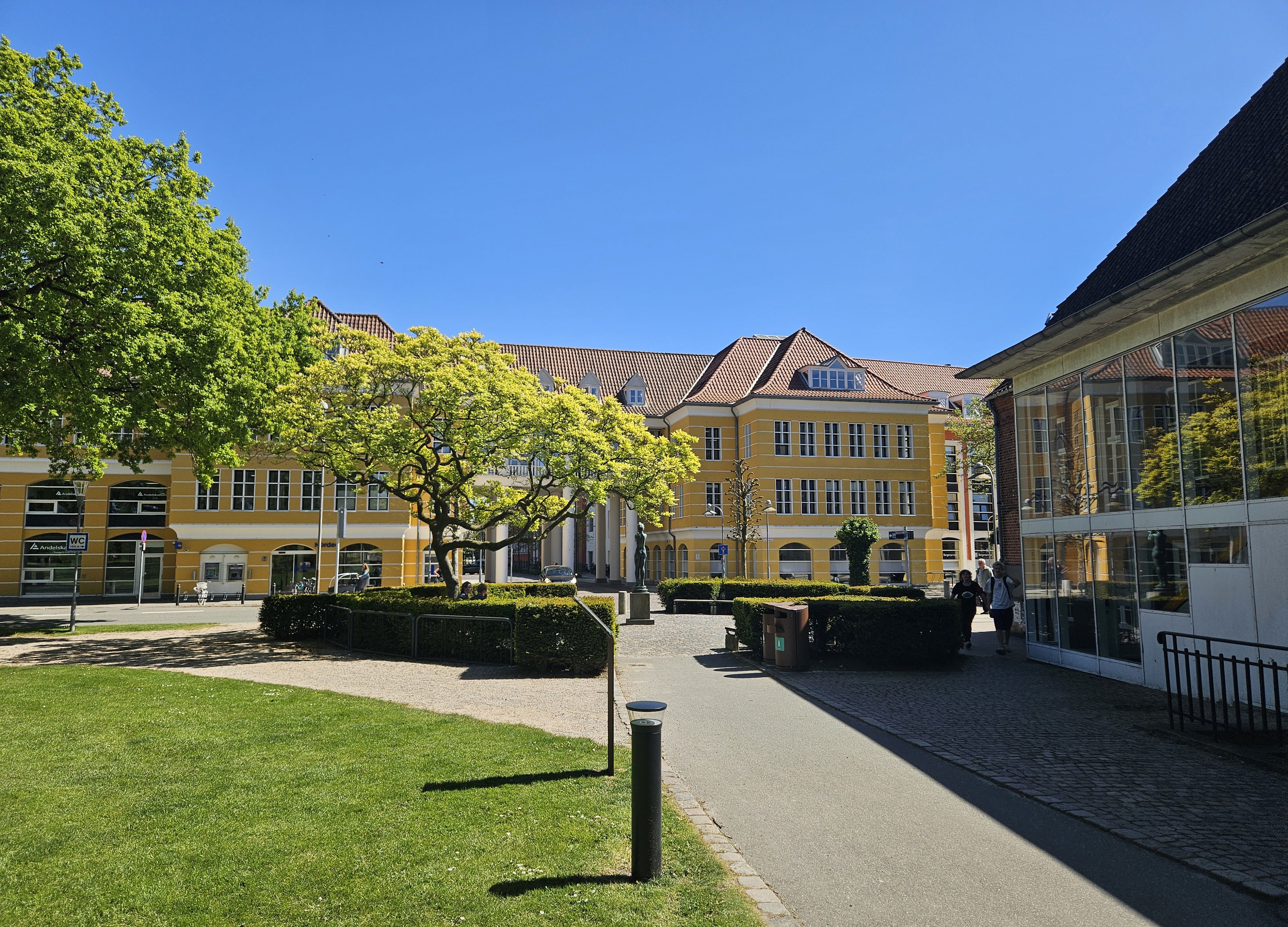 En sti med græs omkring, fører ned til en stor gul bygning med hvide vinduer. Foran bygningen er en plads omkranset af en rund hæk og et stort træ.