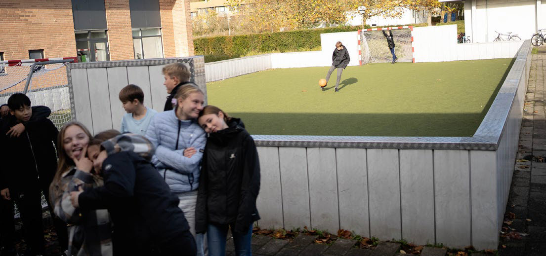 2 drenge spiller fodbold på en lille indhegnet kunststofbane, mens en anden gruppe elever pjatter foran i billedet til ære for fotografen!