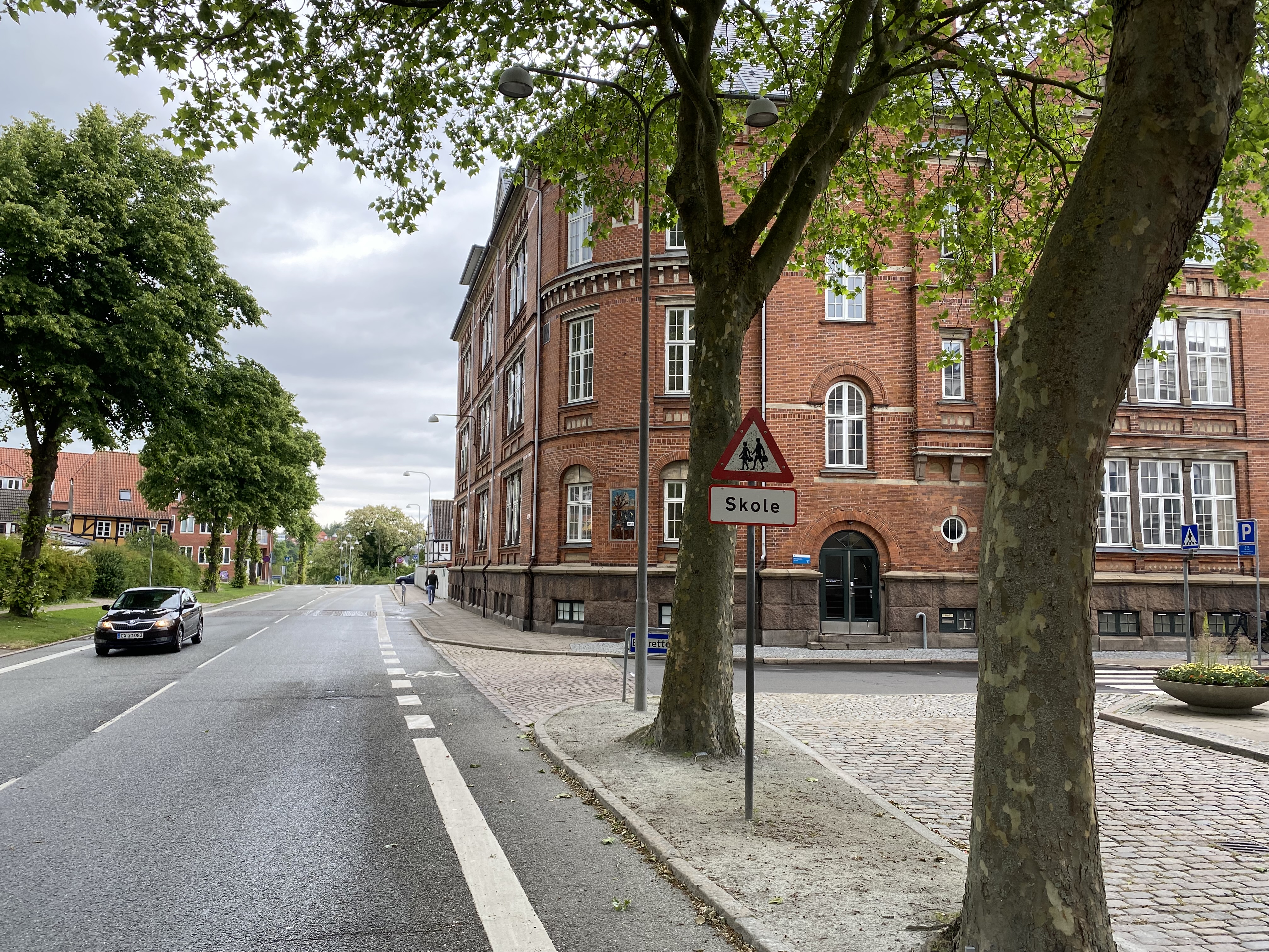 Ørkildskolen afdeling Byen - en gammel rød murstensbygning set fra vejen - udsynet er delvis blokeret med træer i forgrunden og det klassiske trekantede skilt for skole
