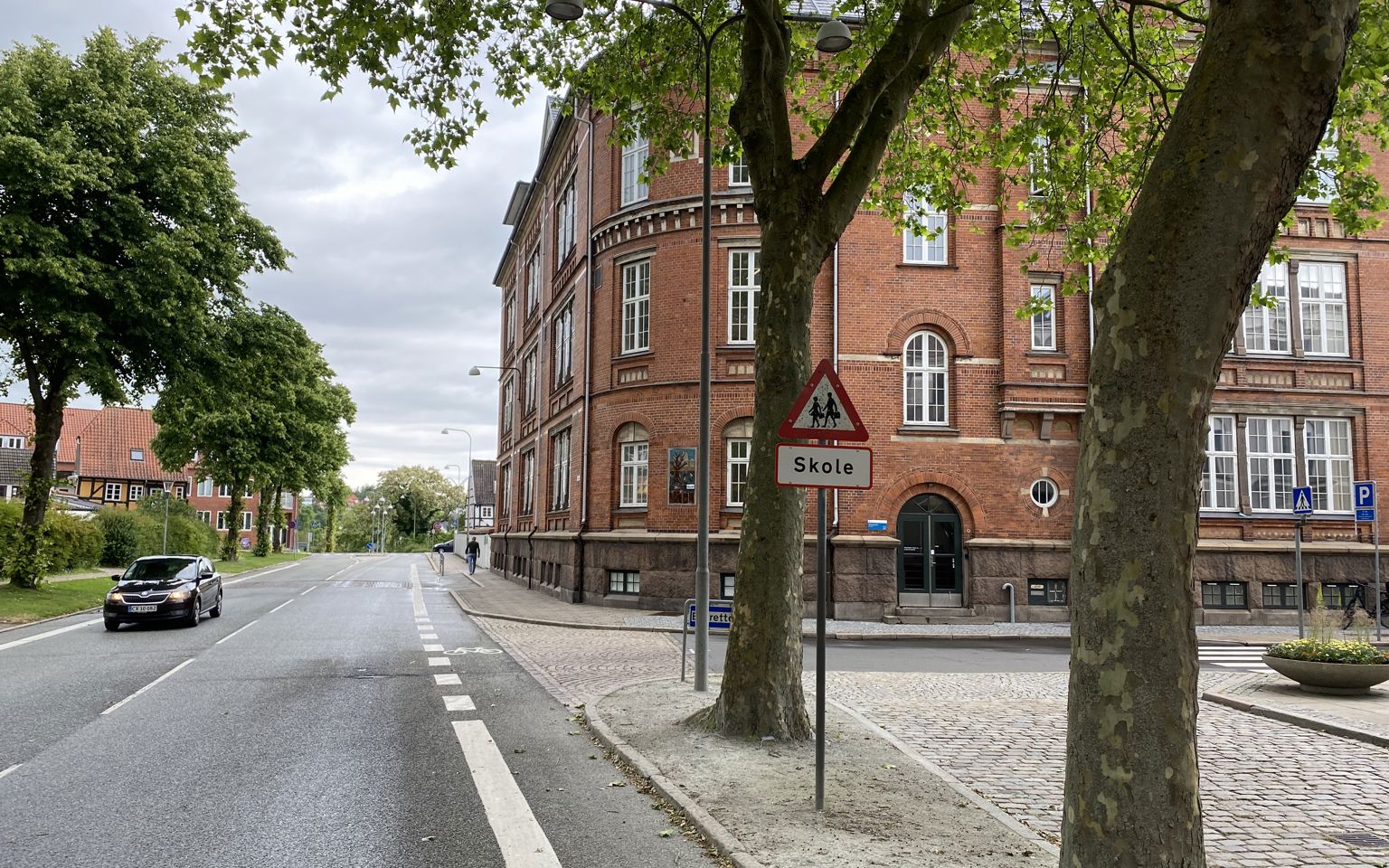 Ørkildskolen afdeling Byen - en gammel rød murstensbygning set fra vejen - udsynet er delvis blokeret med træer i forgrunden og det klassiske trekantede skilt for skole