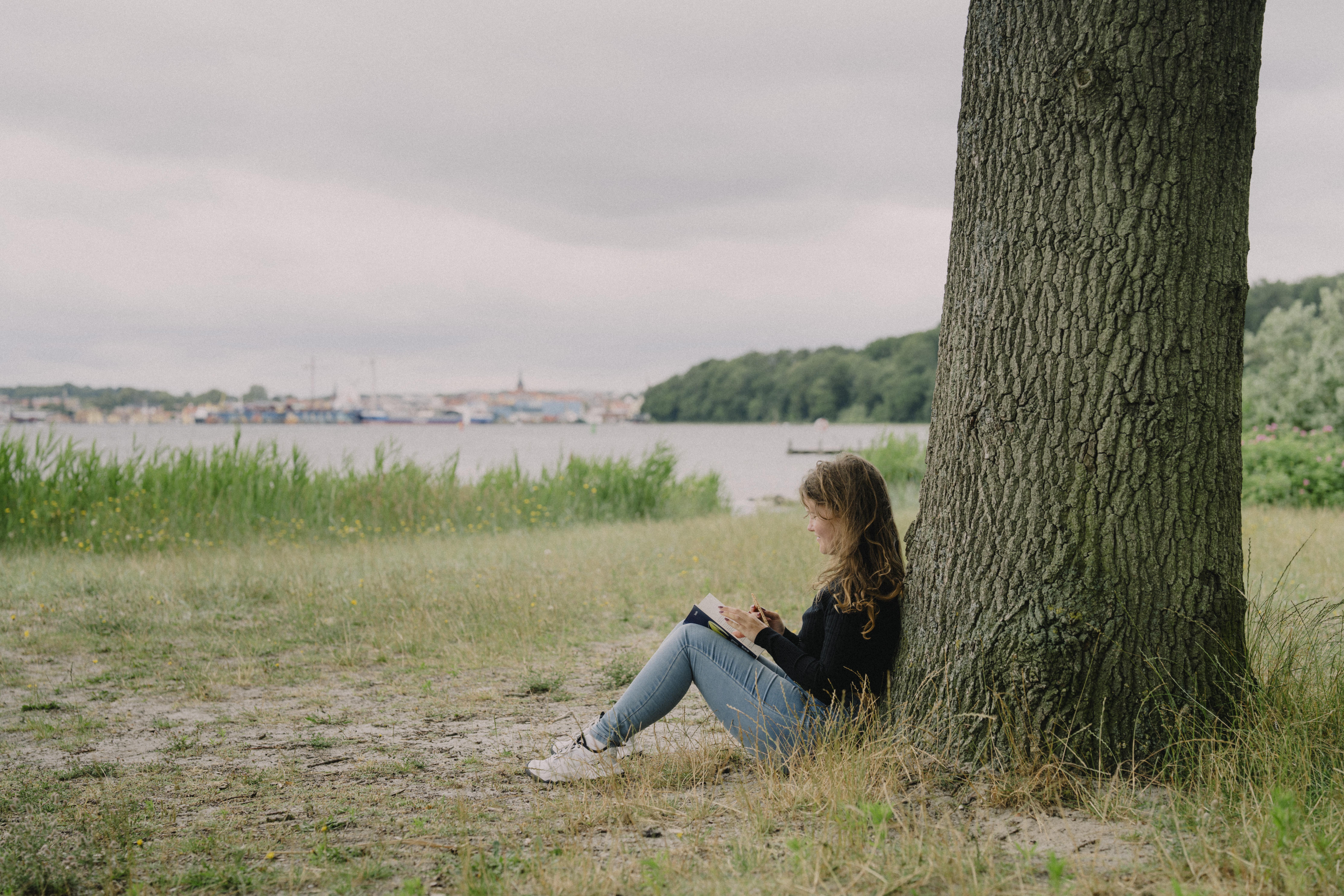 Pige sidder op ad træ og læser bog - i baggrunden Svendborg By set fra vandsiden