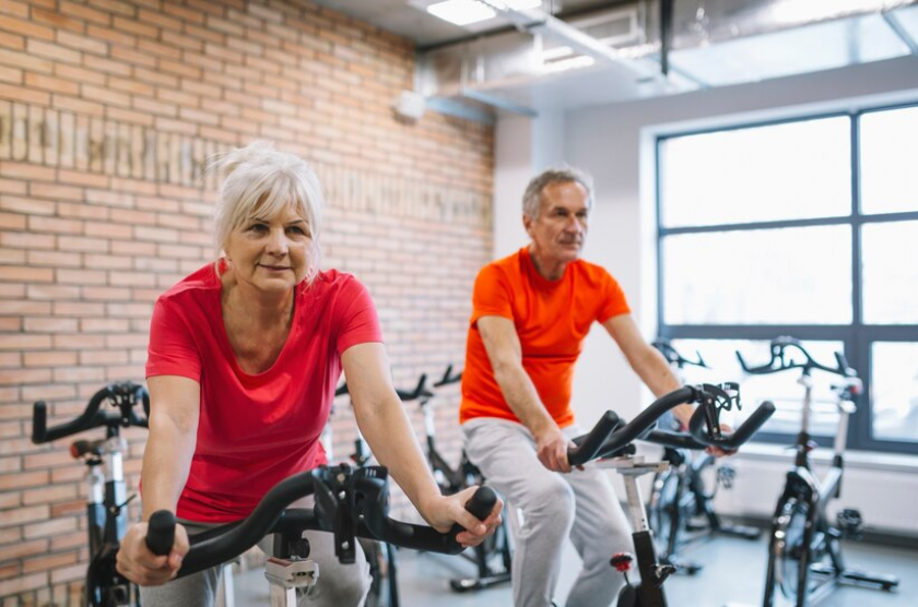 En ældre kvinde sidder på en motionscykel. En ældre mand på en motionscykel ses i baggrunden