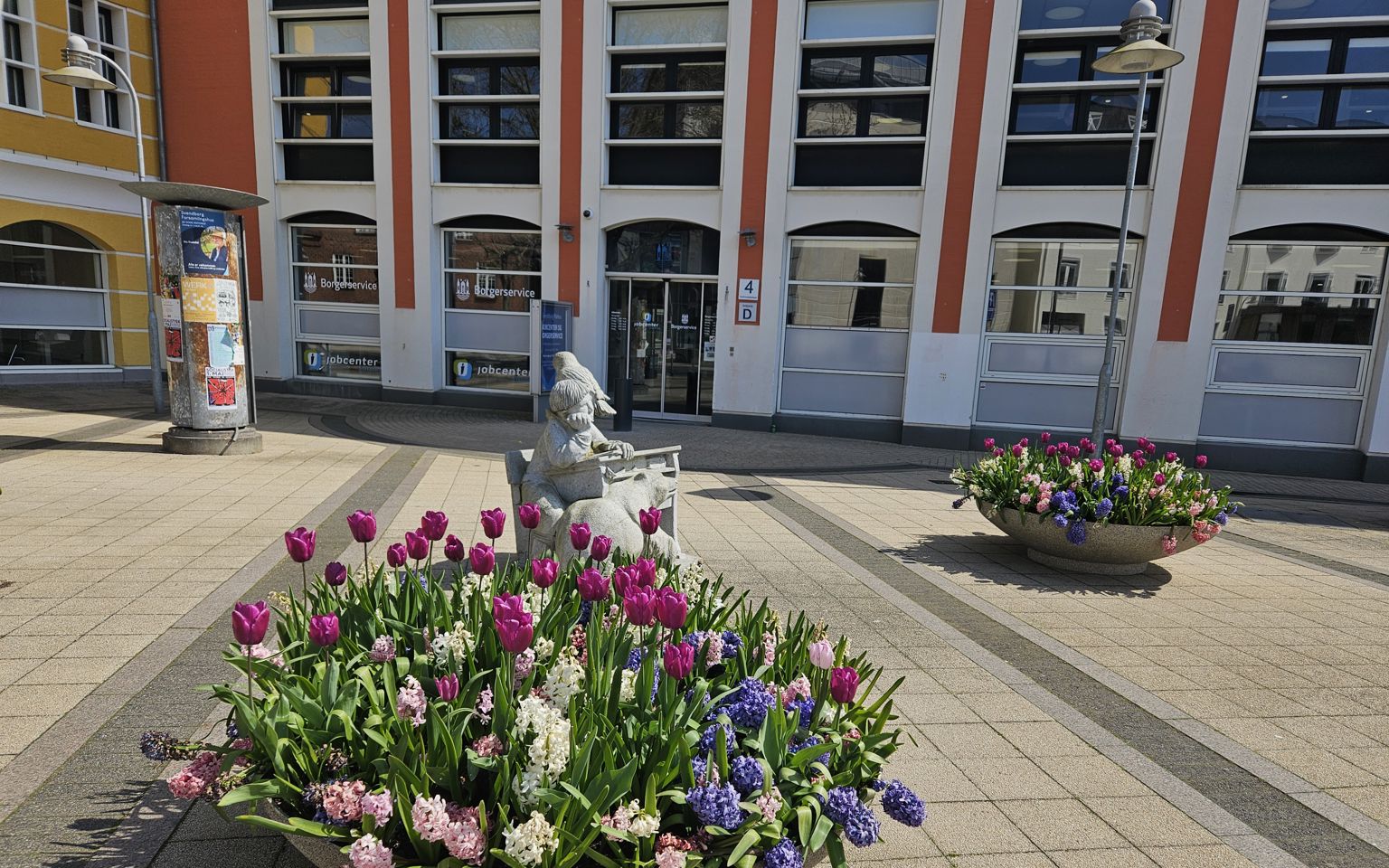 En kumme med forårsblomster foran indgangen til en stor bygning, der er hvid og rød.