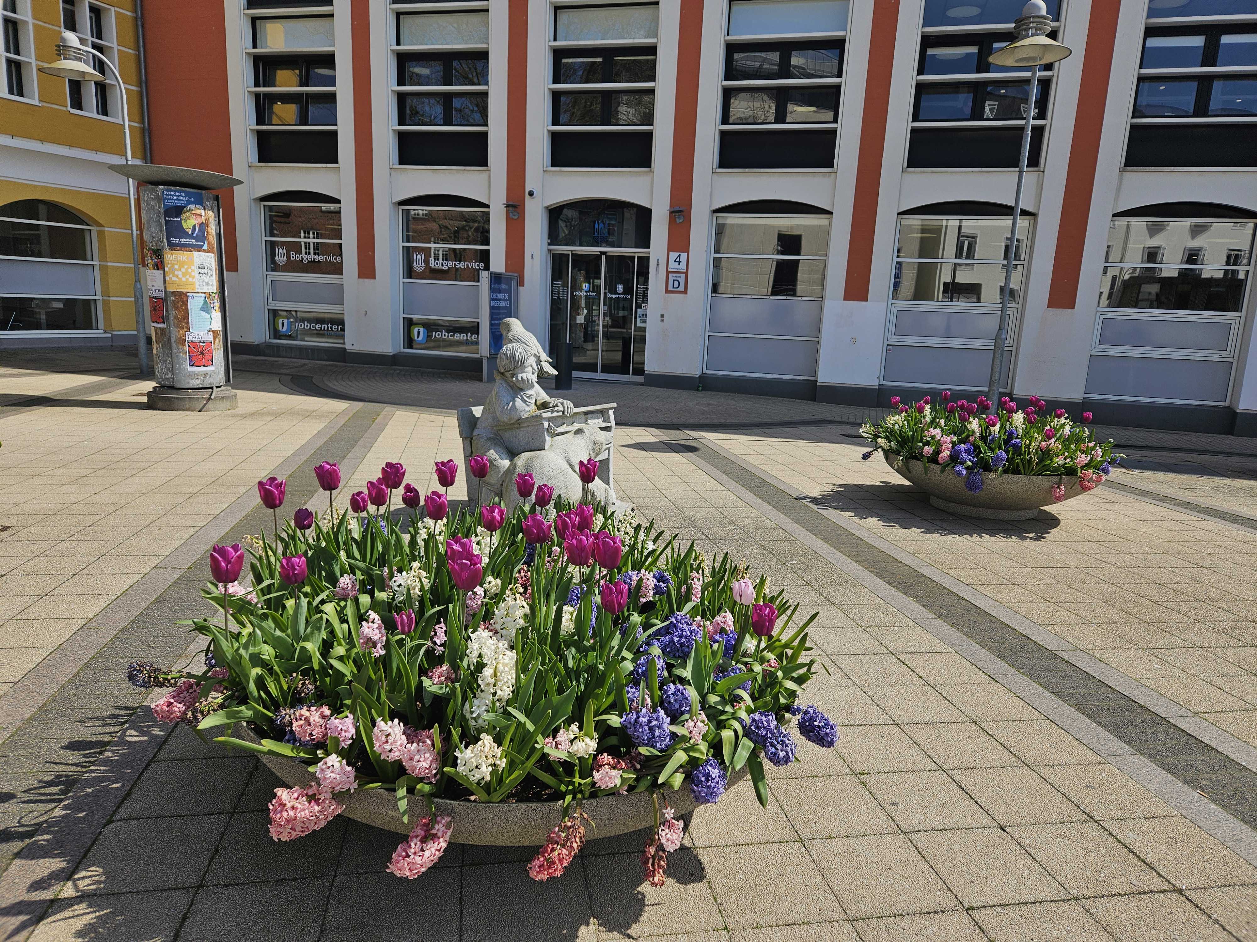 En kumme med forårsblomster foran indgangen til en stor bygning, der er hvid og rød.